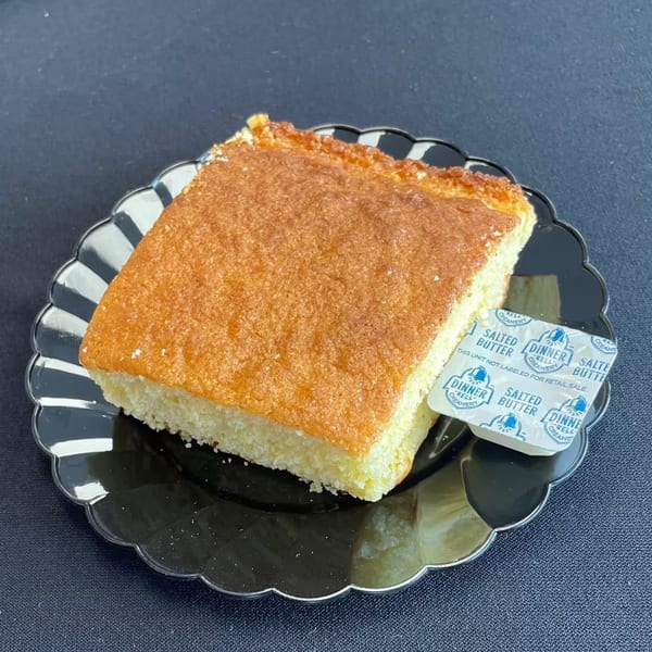 Aunt Berta's Kitchen's golden-brown cornbread slice with butter packets on a black scalloped plate.