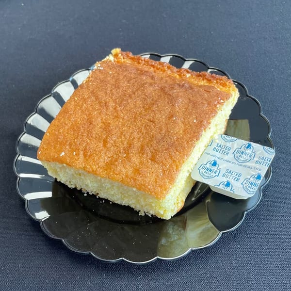 Aunt Berta's Kitchen cornbread slice with a butter packet on a black scalloped plate.