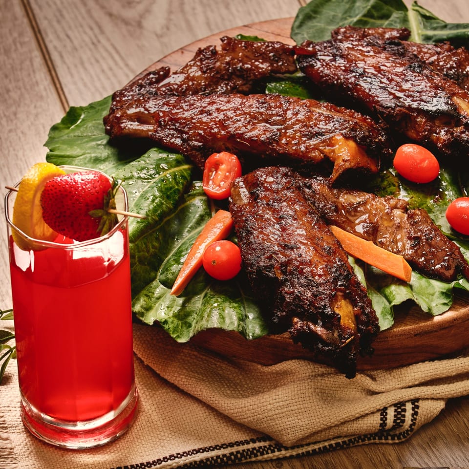 Glazed grilled ribs and vegetables served on leafy greens beside a red fruit drink garnished with strawberry and orange.