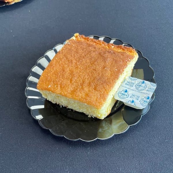 A slice of golden cornbread with a packet of salted butter on a black scalloped plate.