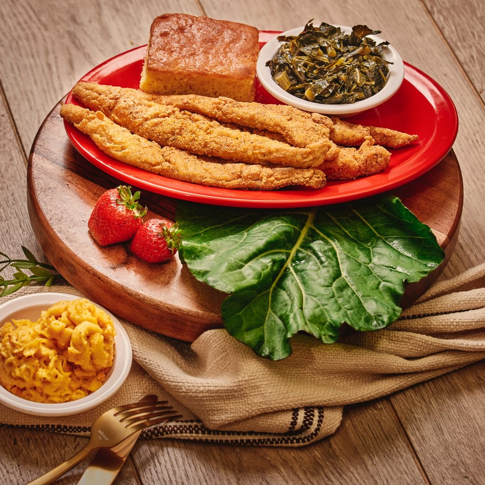 Crispy fried fish, cornbread, collard greens, strawberries, and macaroni and cheese arranged on a red platter.