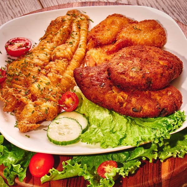 A white plate holds fried fish, shrimp, and crab cakes, garnished with lettuce, tomatoes, and cucumbers.