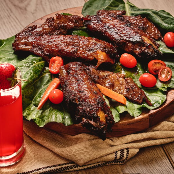 Glazed ribs on a wooden board with green leaves, cherry tomatoes, carrots, and a red drink.